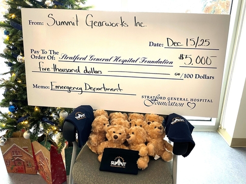 A large ceremonial cheque for $5,000 from Summit Gearworks Inc. to the Stratford General Hospital Foundation is displayed in front of a decorated Christmas tree. In front of the cheque are teddy bears, toques, and shirts donated to the Emergency Department to provide comfort items for patients.
